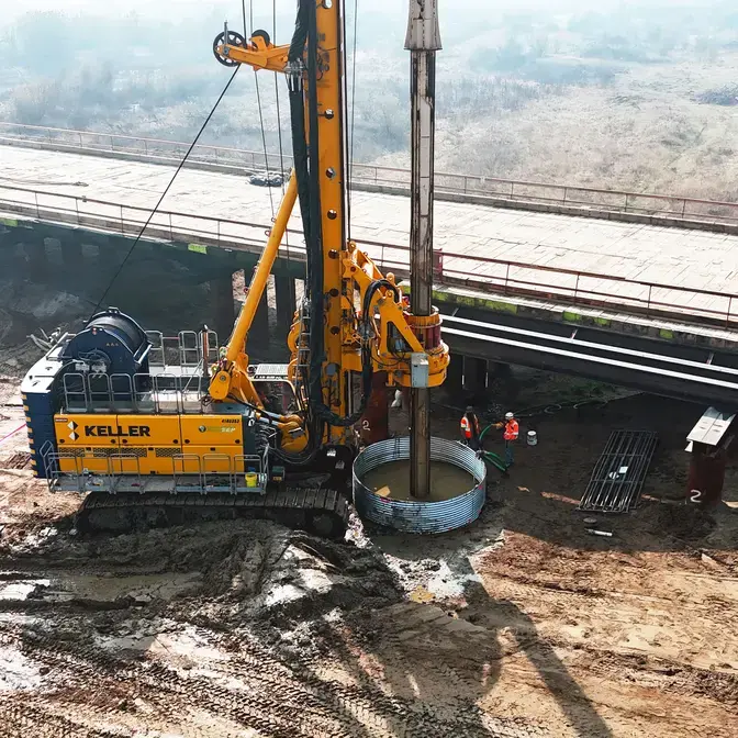 Yellow Keller drilling rig operating beneath an elevated bridge, constructing a drilled shaft while crew members work nearby on a construction site.